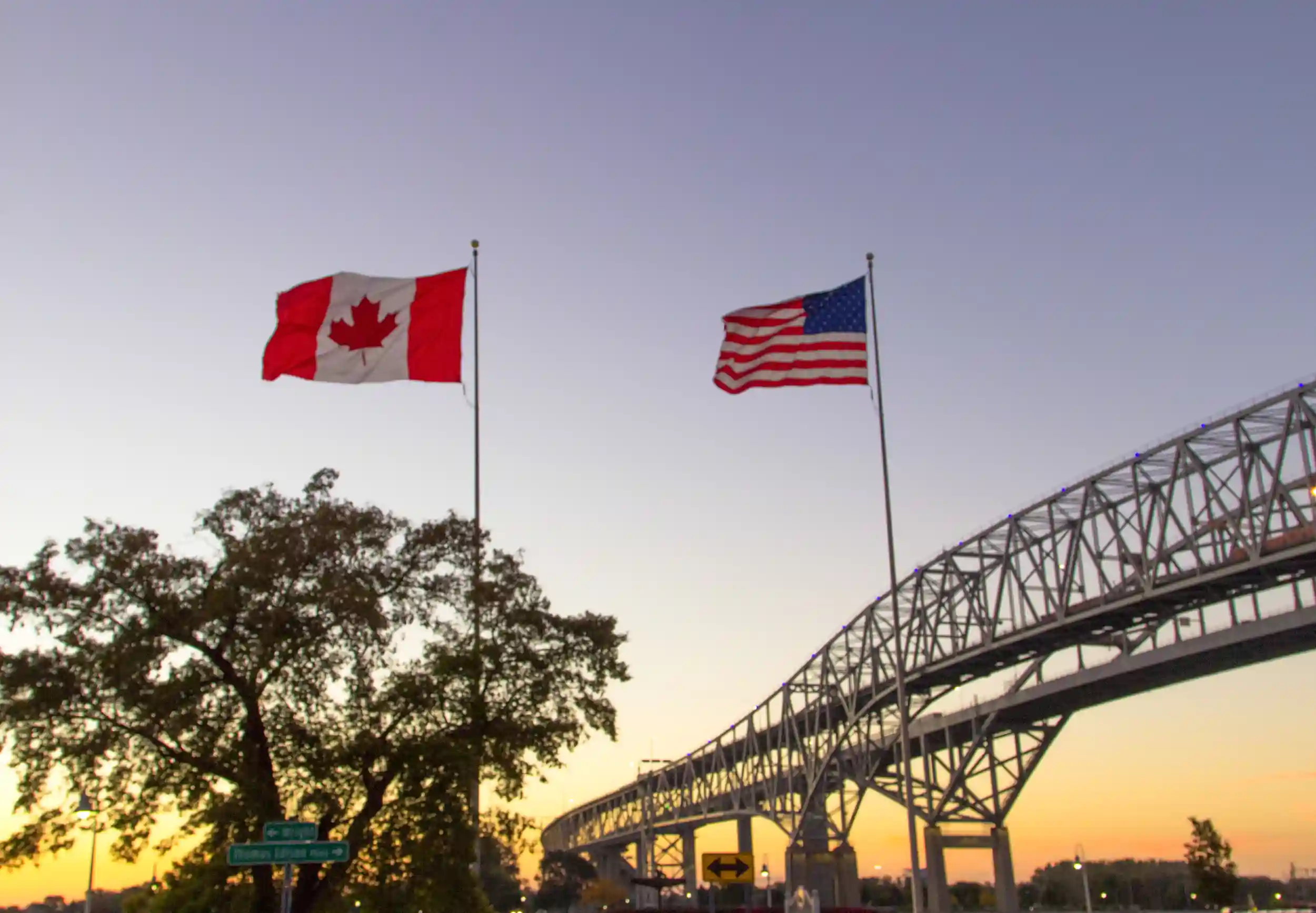 Canada and U.S. flags wave in front of a bridge at sunset.