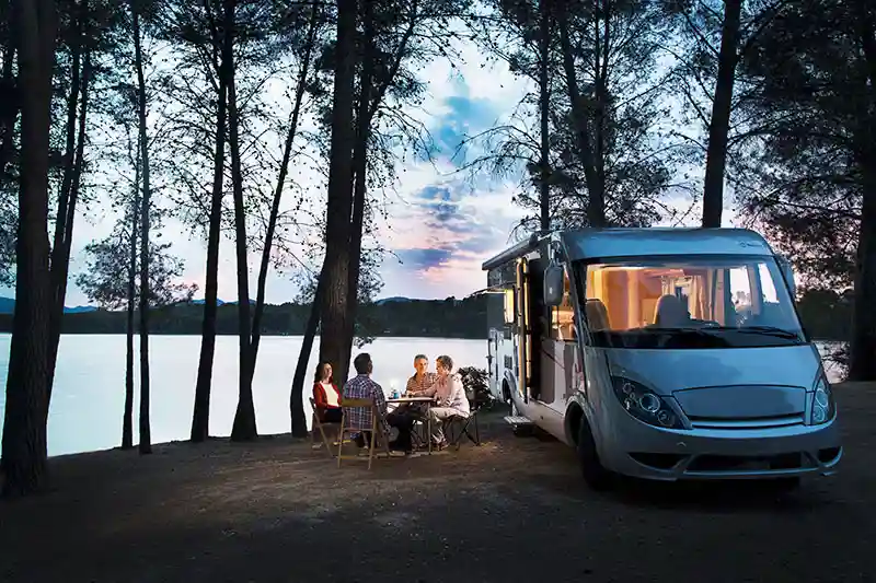 A group of people converse at a lakeside campground next to a large recreational vehicle.
